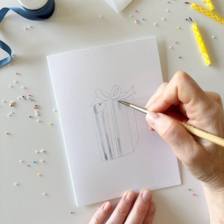 Hand drawing a gift box with a bow on a white card using a paintbrush.