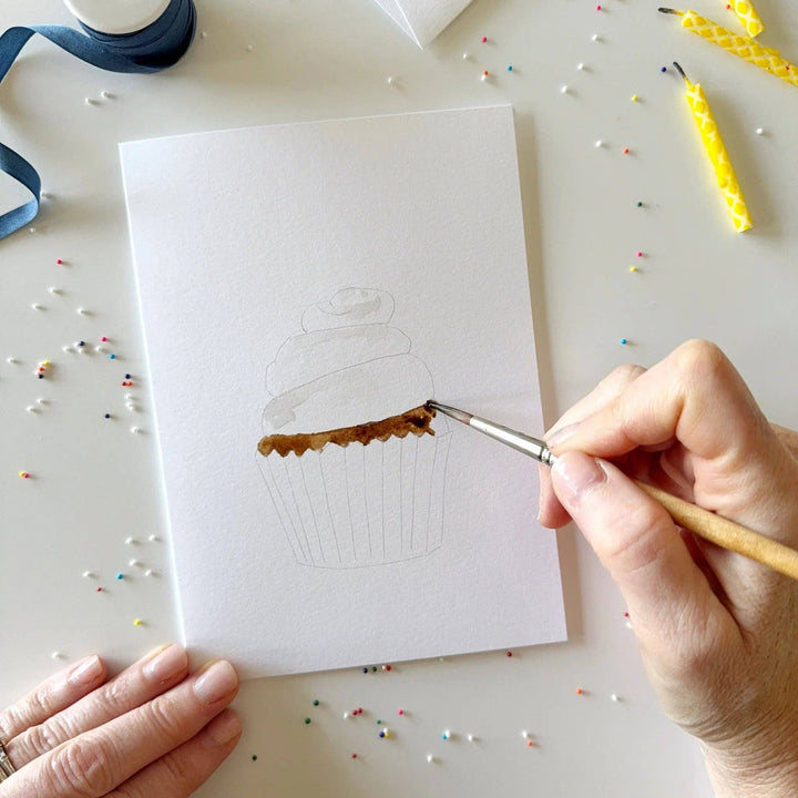 Hand painting a cupcake design on a card with a brush, surrounded by colorful sprinkles and crafting supplies.