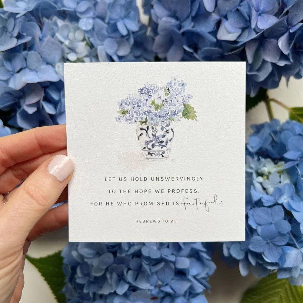 Hand holding a card with a floral illustration and Bible verse text, surrounded by blue hydrangeas.
