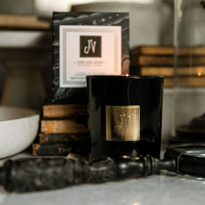 Black candle with gold logo on a surface with books and a bowl in the background