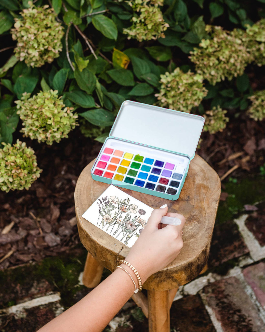 Person painting with watercolors on a small stool outdoors, surrounded by greenery.