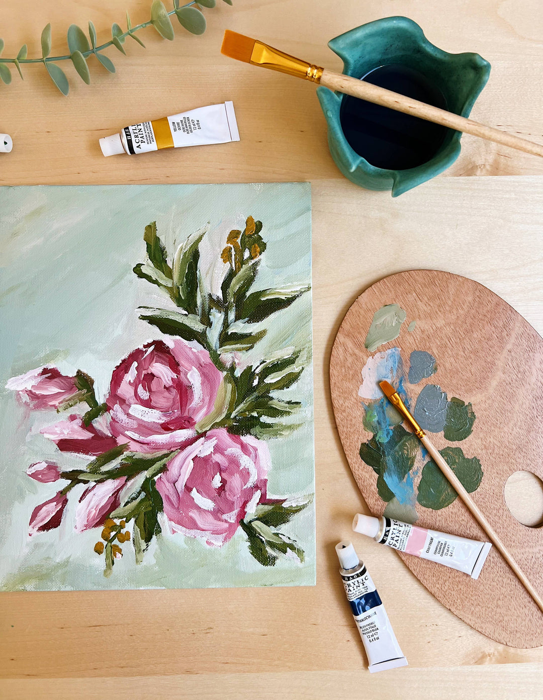 Floral painting on a wooden surface with art supplies including paint tubes, a brush, and a palette.