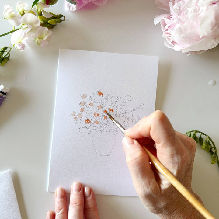 Person painting a floral design on a card with flowers around