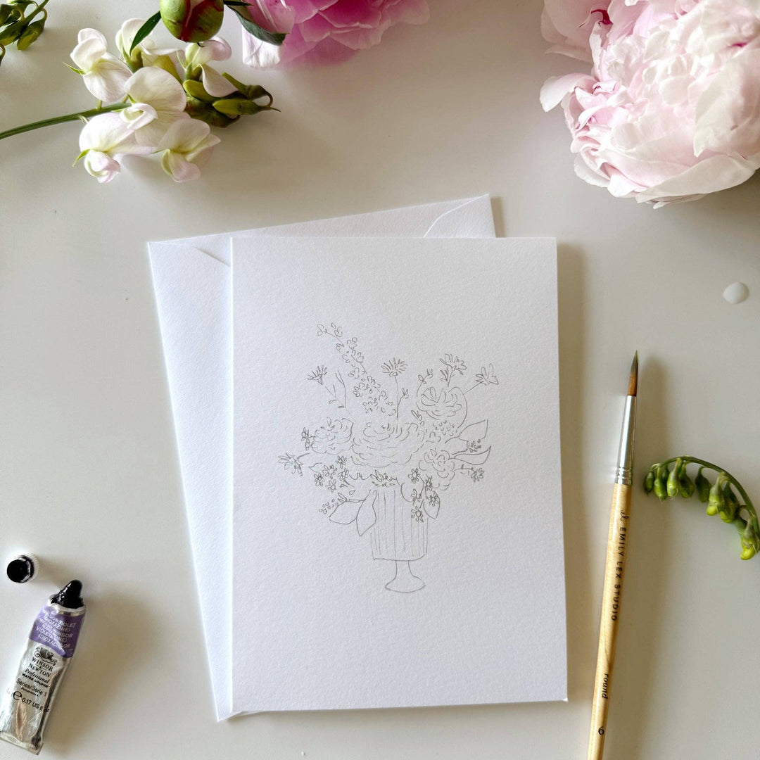 White card with floral design on a white surface with flowers and a paintbrush.