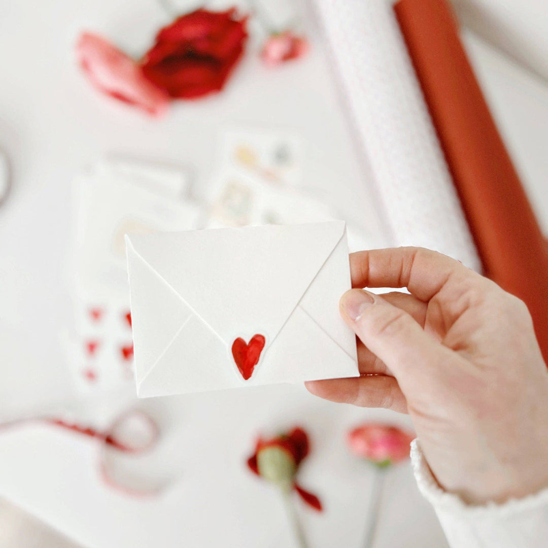 Hand holding a small white envelope with a red heart on a blurred background of flowers.