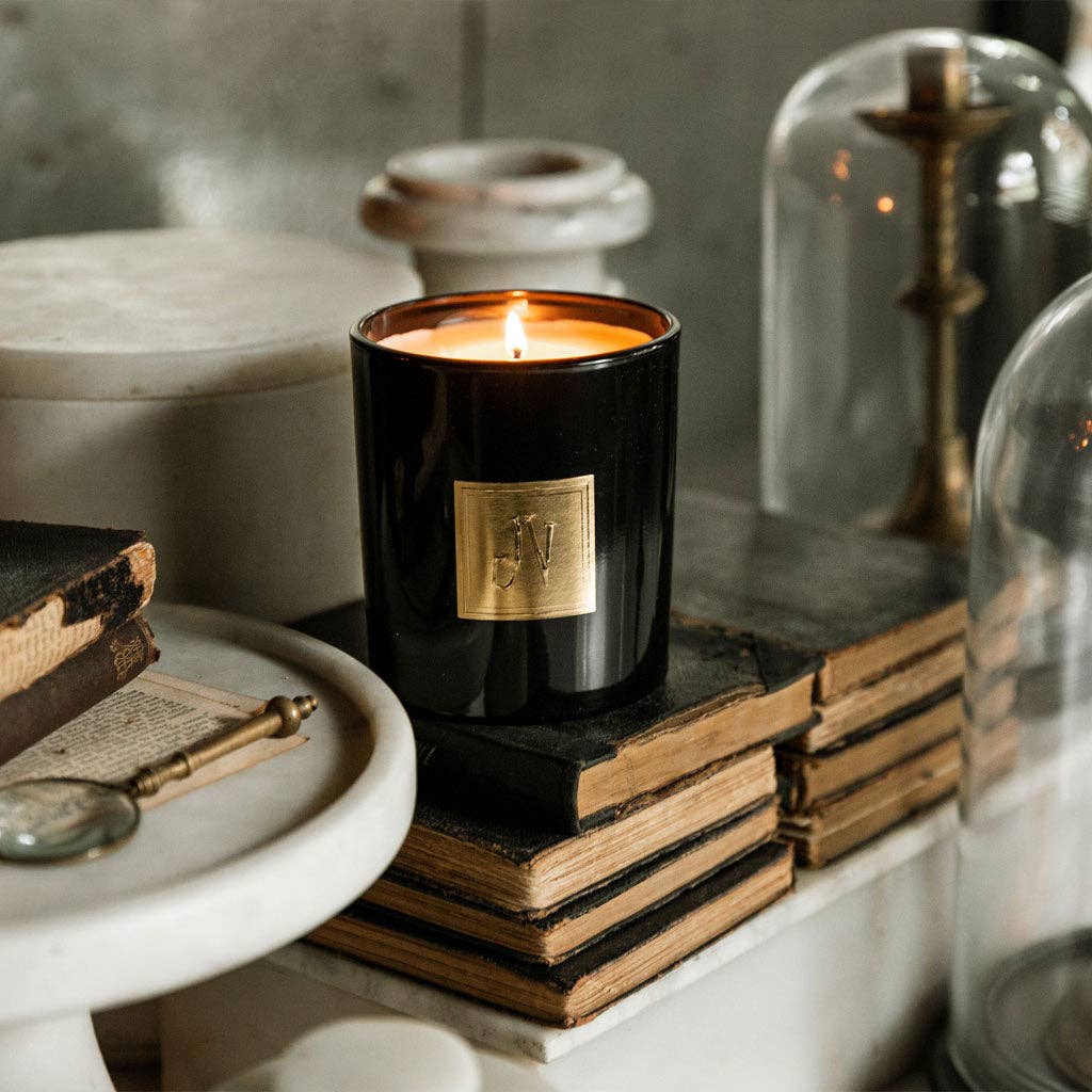Black candle with a gold label on top of old books, surrounded by decorative items.