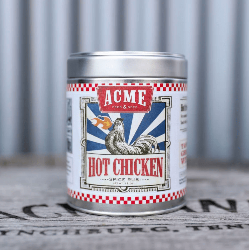 A silver metal tin of Acme Feed & Seed Hot Chicken Spice Rub, featuring a vintage-style label with a rooster and red, white, and blue graphics, set against a blurred gray background.