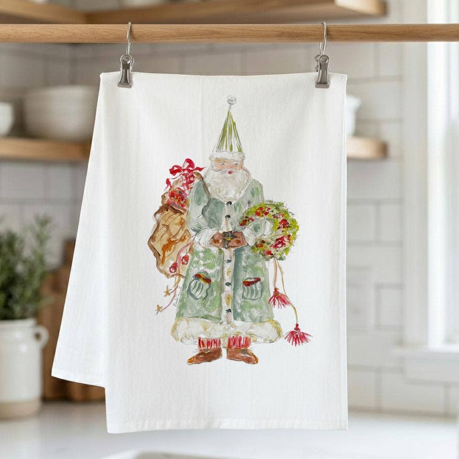 A white kitchen tea towel hanging from a wooden rack with metal clips, featuring a vintage-style illustration of a Santa Claus figure in a green outfit holding a wreath and sack.