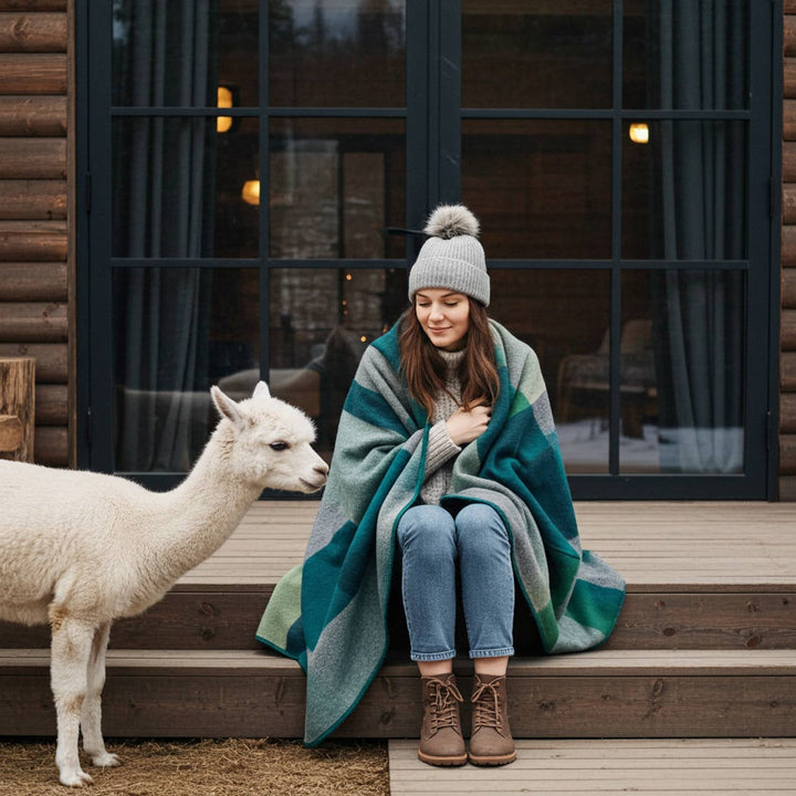 Woman wrapped in a blanket sitting on steps next to an alpaca in front of a log cabin.