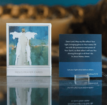 A boxed set of men's prayer cards displayed on a reflective surface. One card in a white frame shows an angel figure against a blue and green background, and another blue prayer card stands beside it with printed text and a small clear stand.