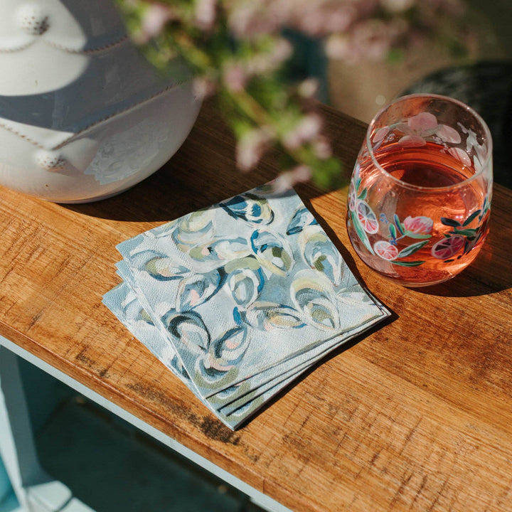 Stack of oyster print napkins on a wooden surface with a glass of pink drink in the background.