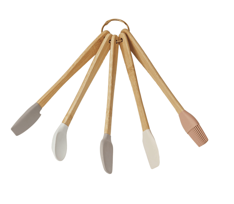 A set of five kitchen utensils with light wood handles and silicone heads in gray, white, and blush pink, hanging from a brass ring on a white background.
