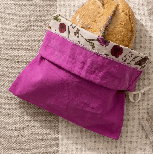 A bright magenta reusable bread bag made of woven fabric, shown holding a loaf of bread. The bag features a floral printed inner lining with the text 'BEE'S WEAR' and a white drawstring closure, set against a neutral woven fabric background.