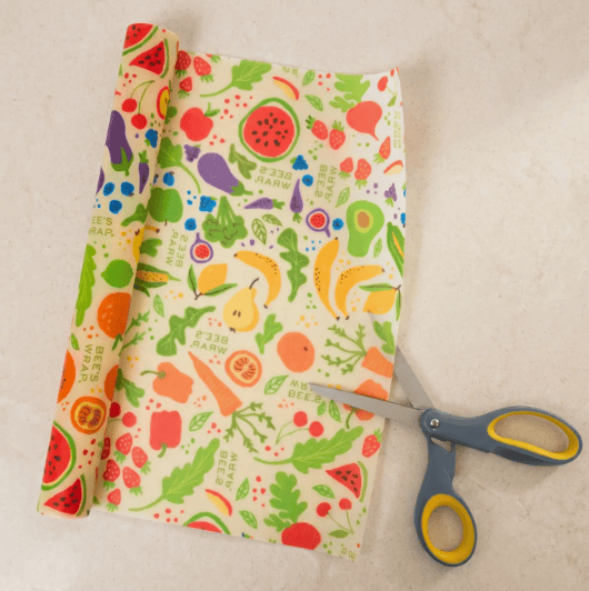 A roll of wax coated fabric with a colorful fruit and vegetable print is laid out on a light beige surface, with a pair of gray and yellow scissors cutting the fabric.