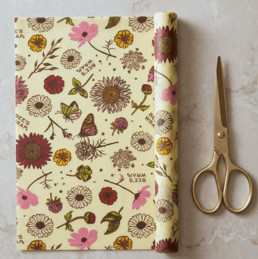 A roll of wax coated food wrap paper with a vintage botanical pattern featuring pink, red, yellow, and green flowers on a cream background, shown next to a pair of gold-handled scissors on a light stone surface.