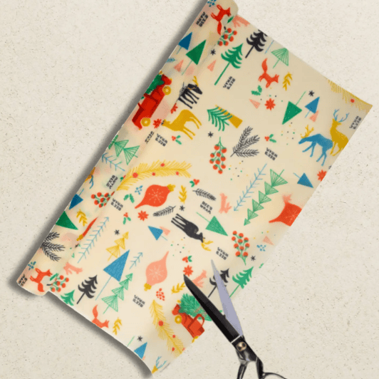 A roll of wax coated food wrap paper  with a colorful holiday woodland animal and tree pattern on a cream background, being cut with black-handled scissors on a light textured surface.