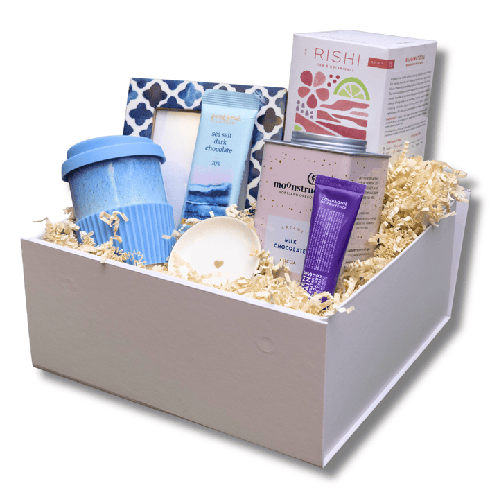 A curated wellness gift box containing a blue travel mug, a patterned soap dish, a blue bottle, a purple tube, a tin, and a Rishi box, all arranged in shredded paper inside a white box.