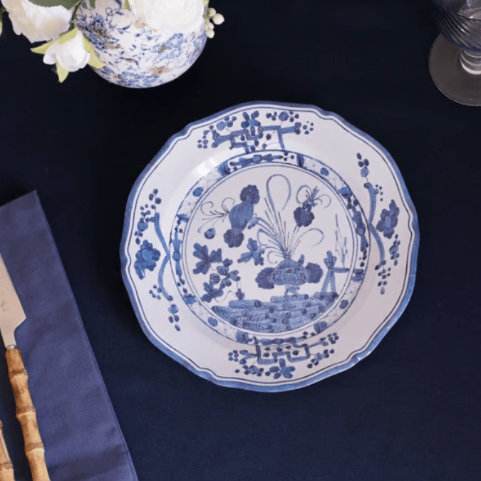 A round white melamine plate with blue floral and landscape illustrations, styled on a dark navy tablecloth with a vase of white flowers and a knife partially visible.
