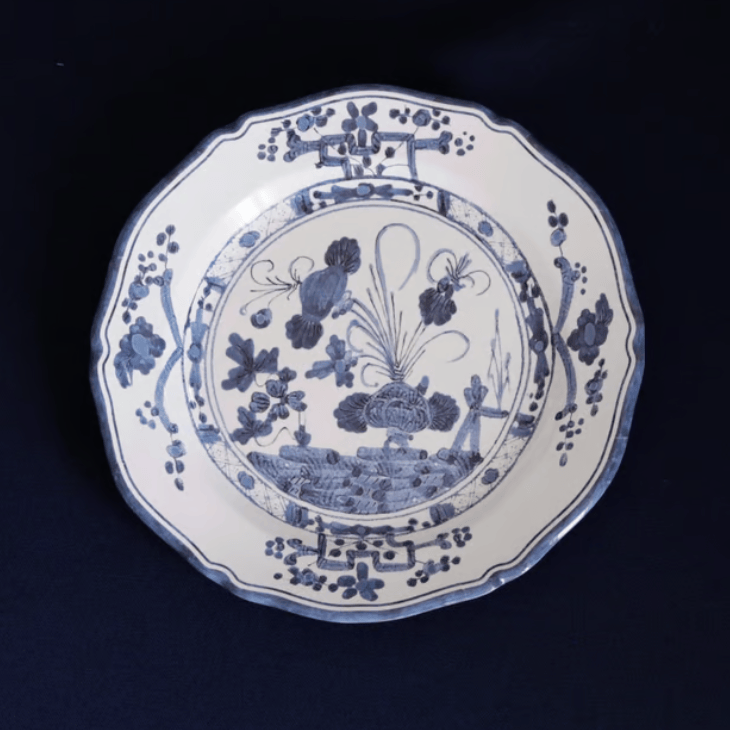 A blue and white melamine plate with a scalloped rim, featuring a detailed blue floral and landscape illustration in the center, set against a dark navy background.