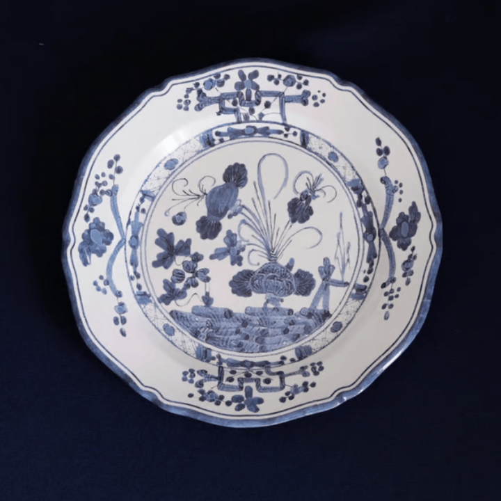 A blue and white melamine plate with a scalloped rim, featuring a detailed blue floral and landscape illustration in the center, set against a dark navy background.