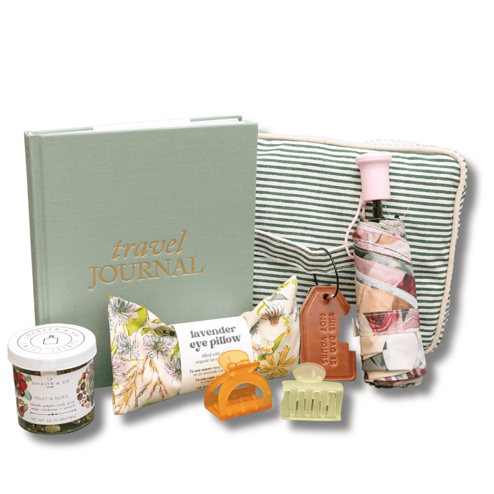 A travel-themed gift set featuring a sage green hardcover journal with gold lettering, a striped zippered pouch, a small jar, a lavender eye pillow, and other travel accessories on a white background.