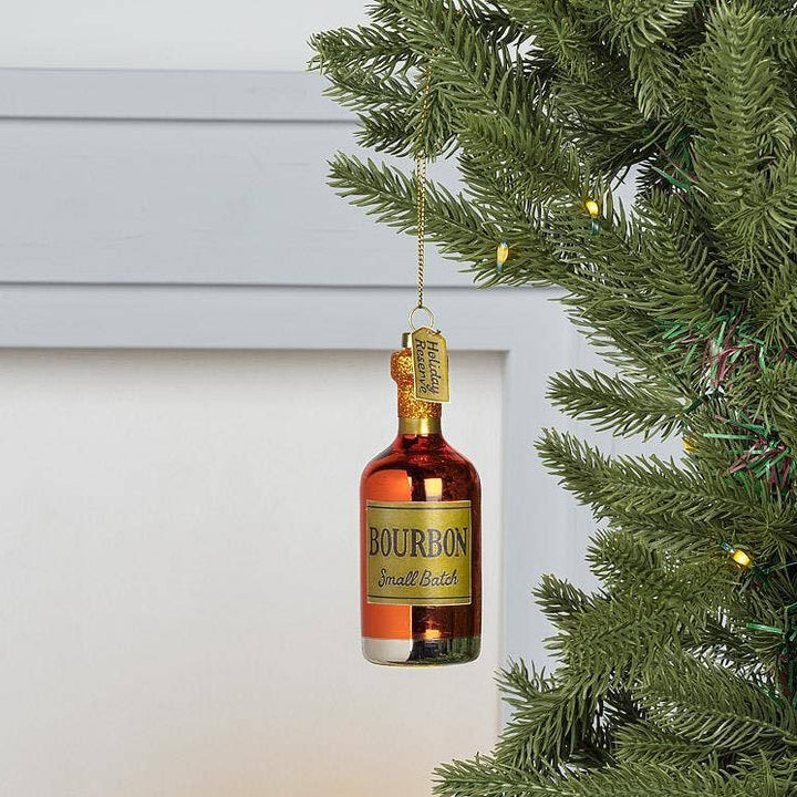 A decorative Christmas ornament shaped like a Bourbon whiskey bottle hangs from a green Christmas tree branch against a white paneled wall.