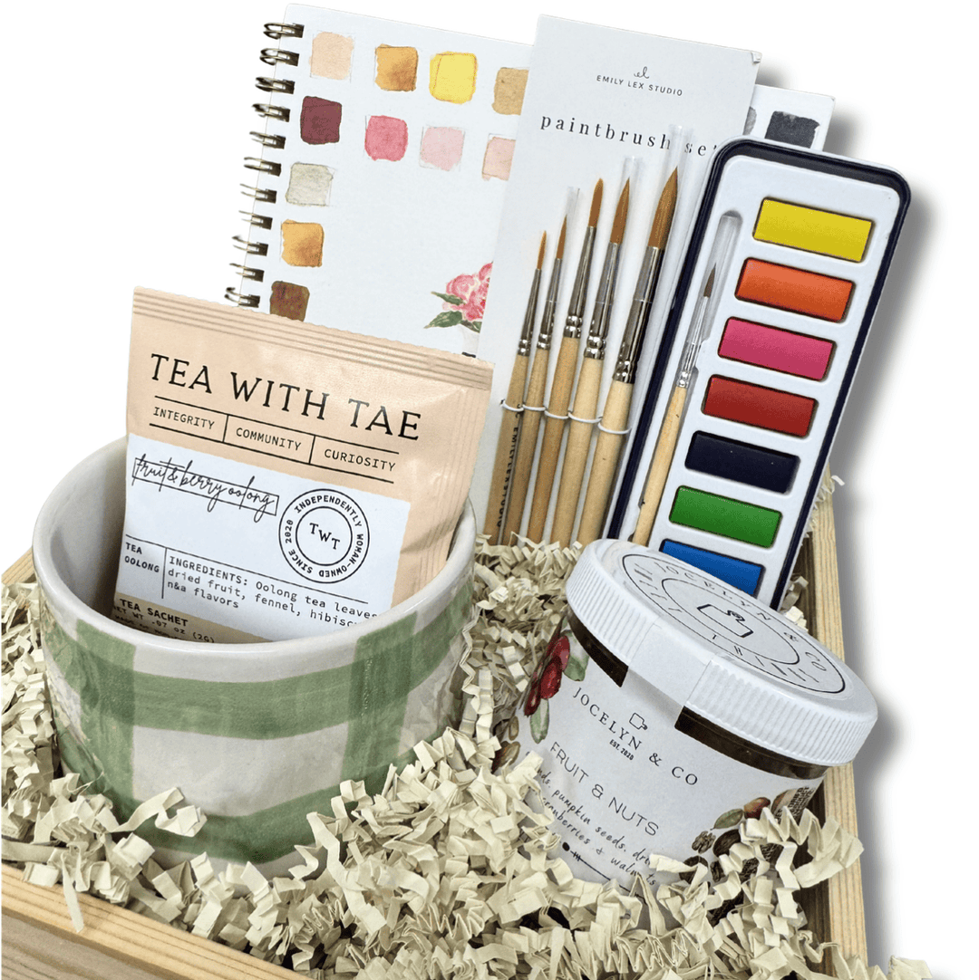 A curated gift basket or tray arrangement on a white background, featuring a green-and-white checkered cup, a tea packet labeled 'TEA WITH TAE', a jar of fruit and nuts from 'The Fruit & Nut Co', a paintbrush set, a watercolor palette, and a swatch notebook.