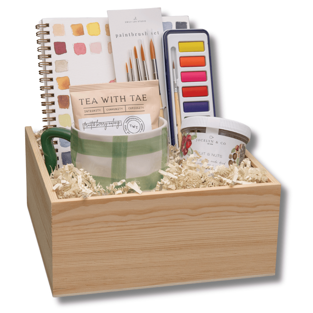 A wooden gift box filled with stationery and art supplies, including a spiral notebook, watercolor paints, a striped mug, and a small jar, set against a plain white background.