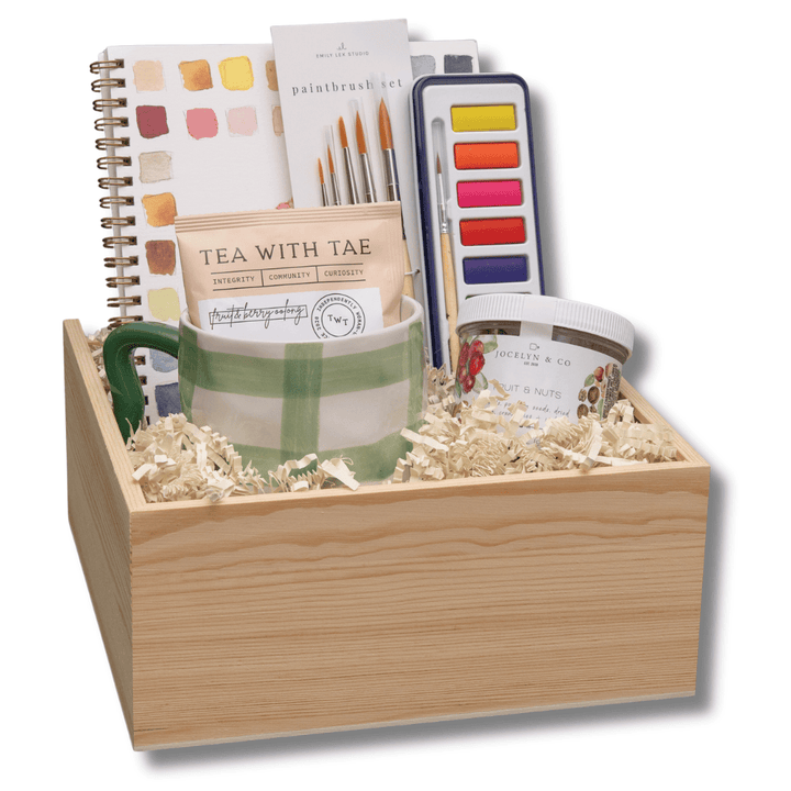 A wooden gift box filled with stationery and art supplies, including a spiral notebook, watercolor paints, a striped mug, and a small jar, set against a plain white background.