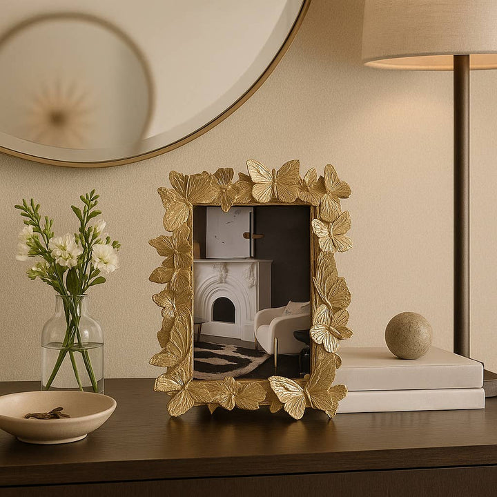 Decorative gold frame with butterfly design on a table, with a vase of flowers and books in the background.