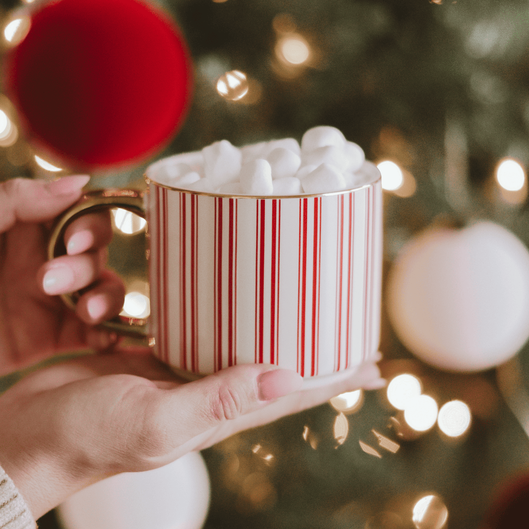 Hand holding a mug with red and white stripes filled with hot chocolate and marshmallows, against a Christmas tree background.