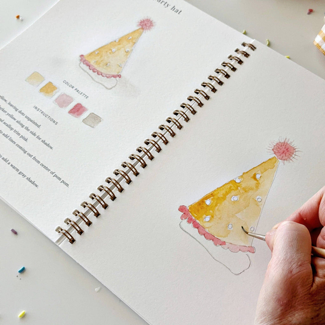 Person painting a party hat illustration in a sketchbook with watercolor techniques.