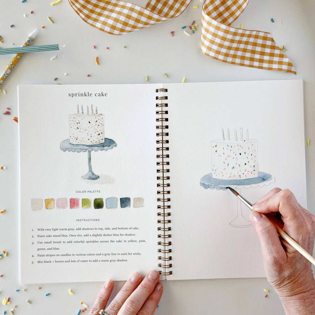A person's hands are shown painting a cake on an open spiral-bound sketchbook. The page features a watercolor reference of a sprinkle cake with a color palette and instructions.