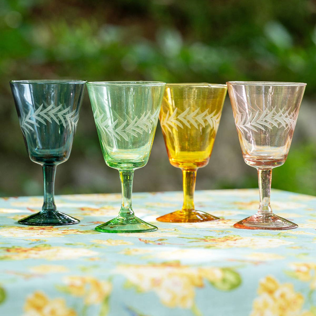 Four etched glass goblets in green, blue, yellow, and pink on a floral tablecloth with a blurred natural background.
