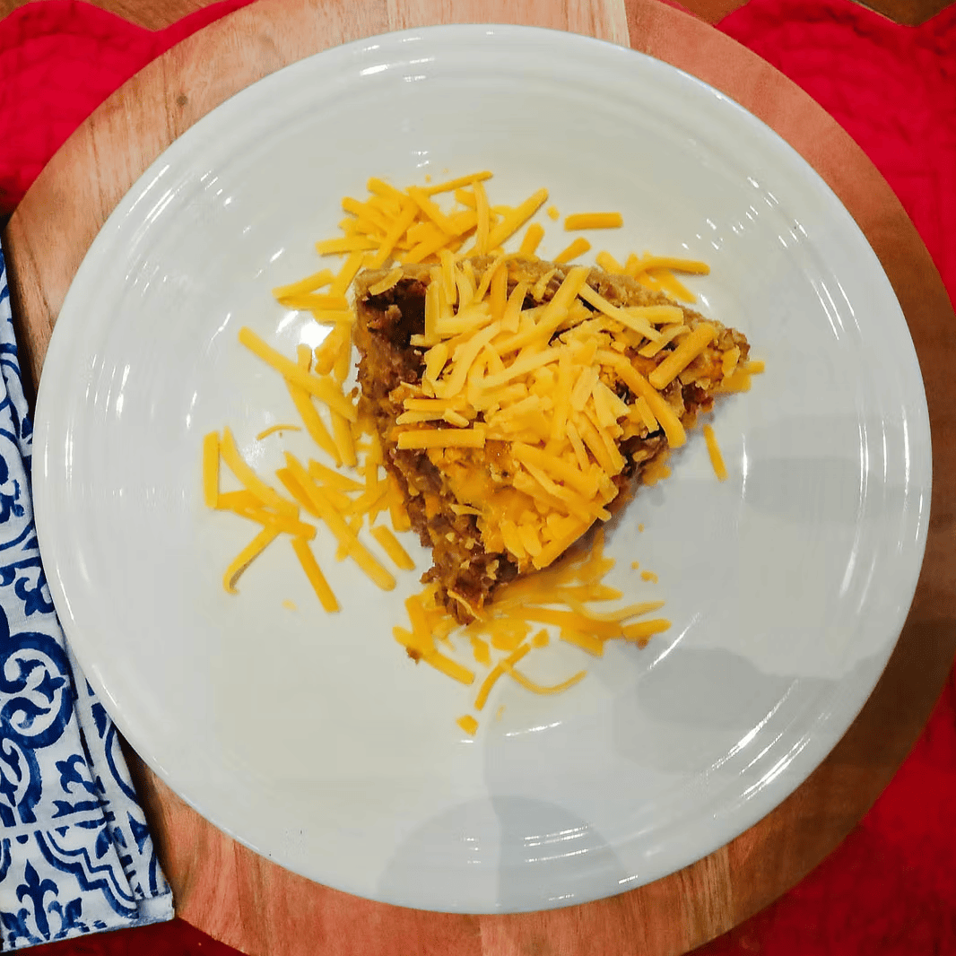 Slice of pie with shredded cheese on a white plate, placed on a wooden table with a red cloth.