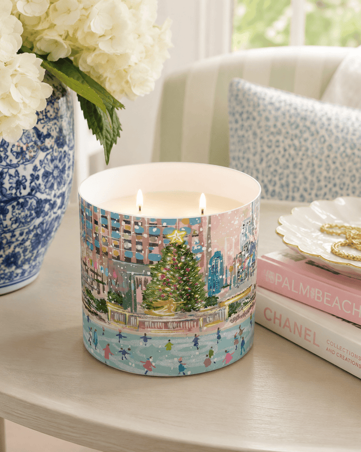 A double-wick candle in a cylindrical tin with a colorful winter city scene printed on the outside, lit with two flames. The candle sits on a light wood table next to a blue-and-white ceramic vase with white flowers, a stack of pink and white books, and a decorative dish with gold jewelry in a softly lit interior setting.