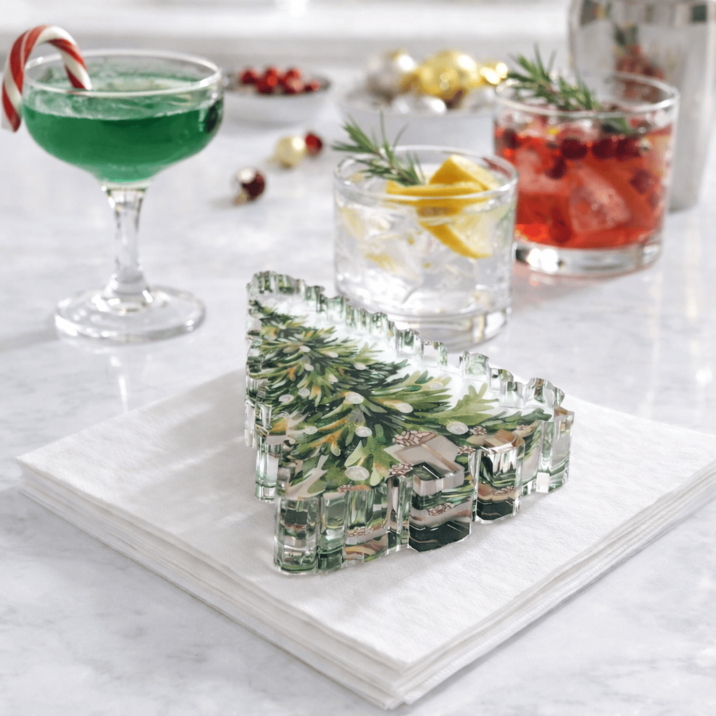 A Christmas tree-shaped clear acrylic napkin weight with a printed green pine tree design, displayed on a stack of white paper napkins on a light marble surface. In the softly blurred background are holiday-themed drinks in glasses and a cocktail glass.