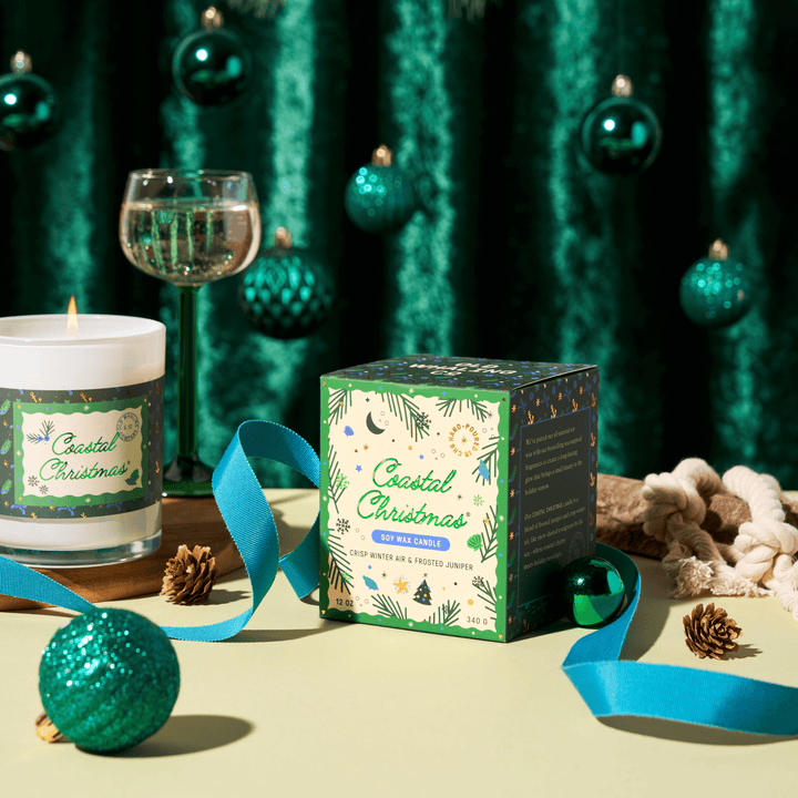 A Coastal Christmas scented candle and its decorative box are styled on a pale yellow surface with teal ornaments, pinecones, a blue ribbon, a wine glass, and a green curtain backdrop.