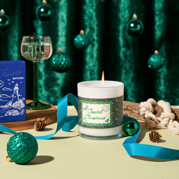 A lit white candle in a clear glass jar with a green and gold holiday label reading 'Coastal Christmas' sits on a pale yellow surface. The scene is styled with teal ribbons, pinecones, a green ornament, a blue Fanbea Mates box, a green stemmed glass, and a dark green velvet curtain backdrop with hanging ornaments.