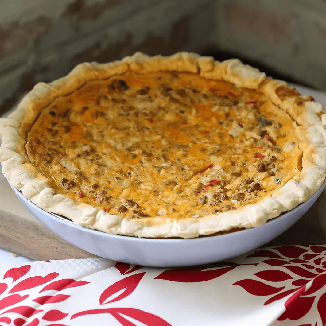 Quiche in a pie dish on a wooden cutting board with a red and white patterned cloth underneath.