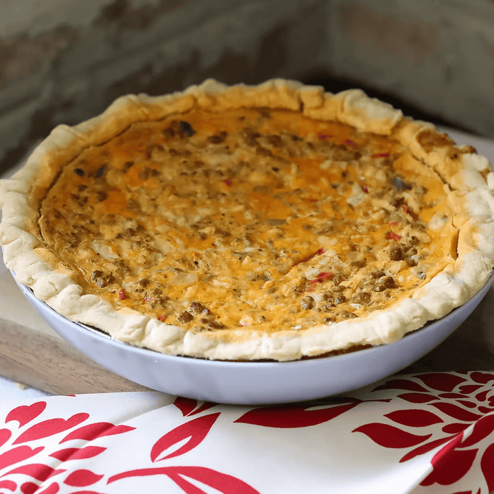 Quiche in a pie dish on a wooden cutting board with a red and white patterned cloth underneath.