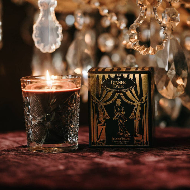 Candle in a glass and 'Dinner Date' card box on a textured surface with blurred decorative lights in the background.