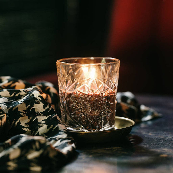 Maroon colored candle in cut glass holder on a dark surface with a patterned cloth.