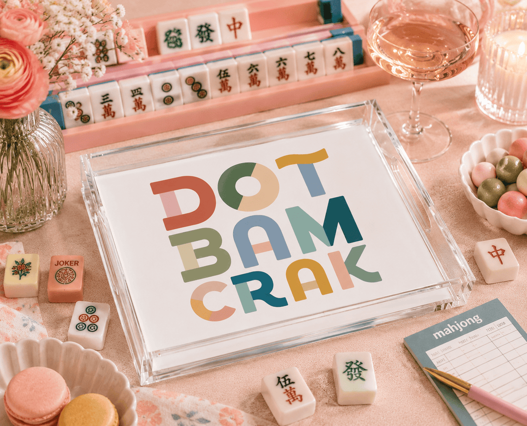 A clear acrylic serving tray with a white insert printed with the phrase “DOT BAM CRAK” in large multicolored letters, styled on a pink tabletop with mahjong tiles, flowers, macarons, a glass of rosé wine, and a notebook.