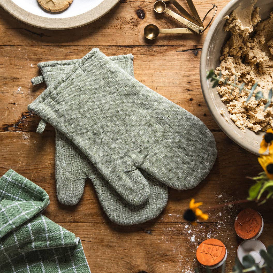Green oven mitts on a wooden table with baking ingredients and tools.