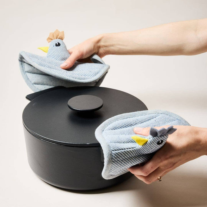Person holding a hen potholders over a black pot on a light background