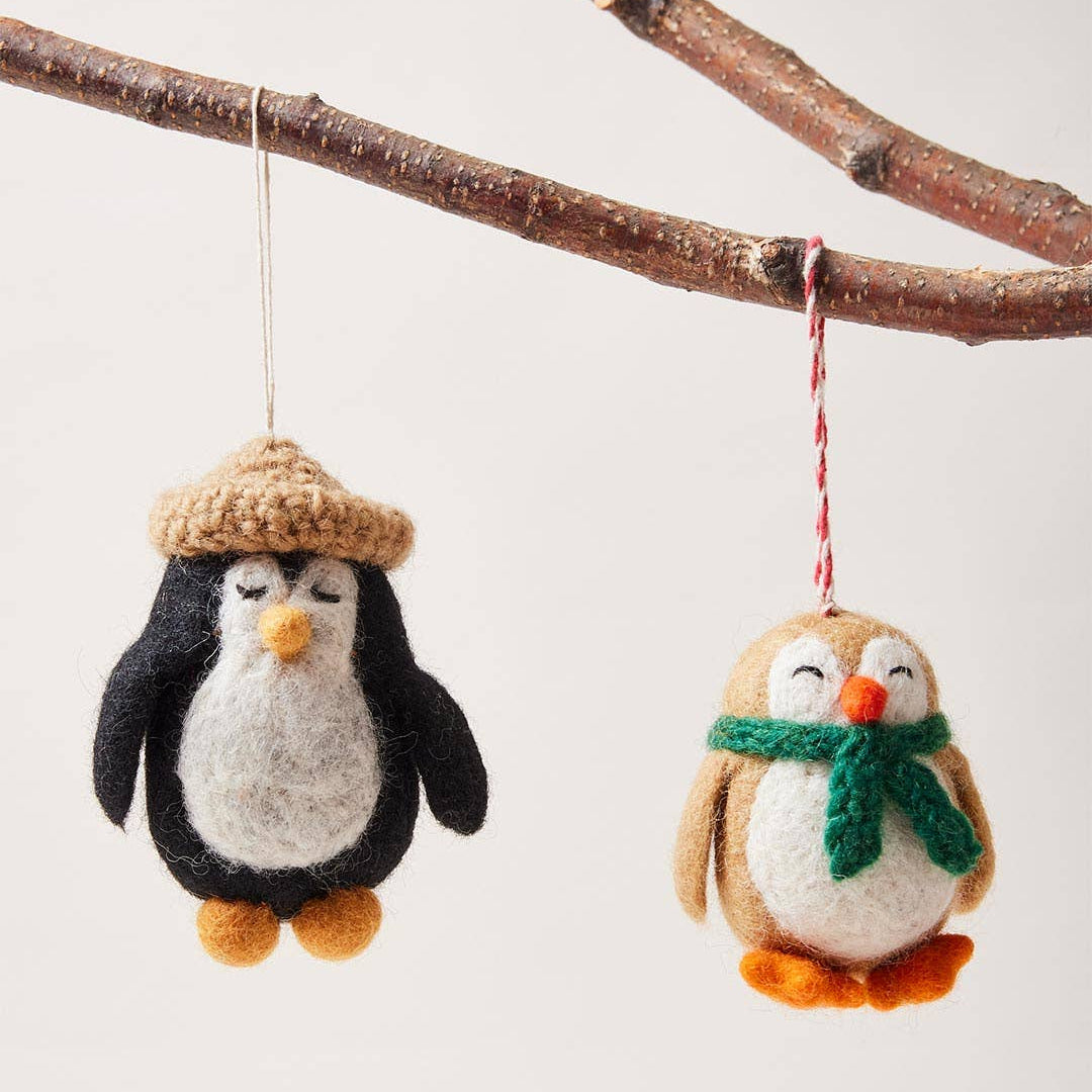 Two felted penguin ornaments with a hat and scarf hanging from a branch.