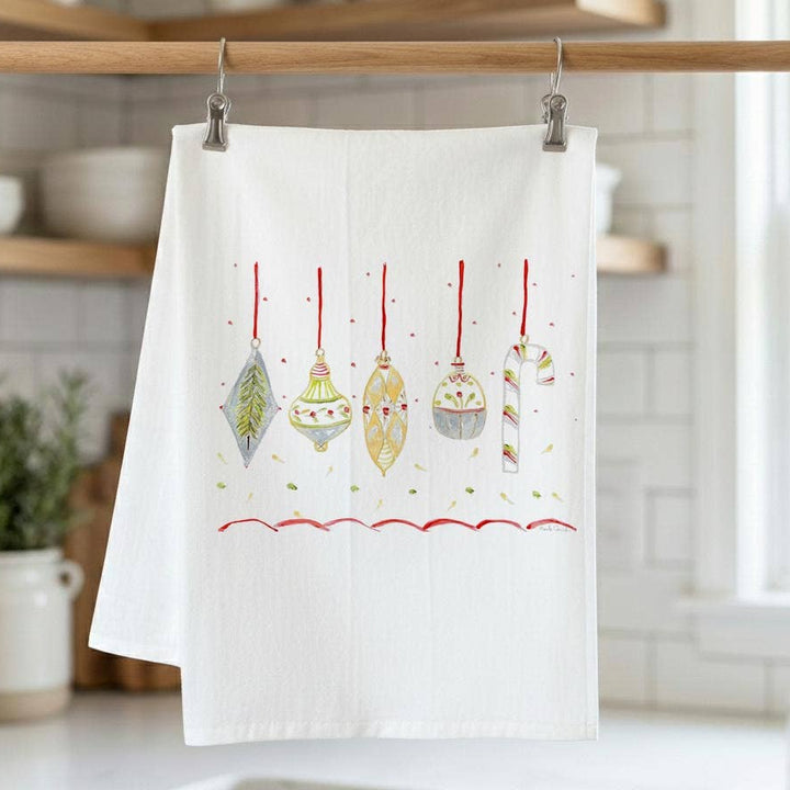 A white kitchen tea towel hanging from a wooden rod with metal clips, featuring a festive illustration of six hanging Christmas ornaments in red, gold, green, and silver. The background shows a softly blurred kitchen interior with white subway tiles, wooden shelves, and a window.