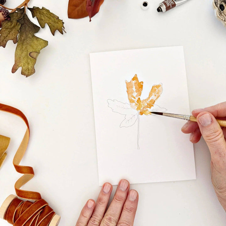 Person painting a leaf design on a card with autumn-themed elements.