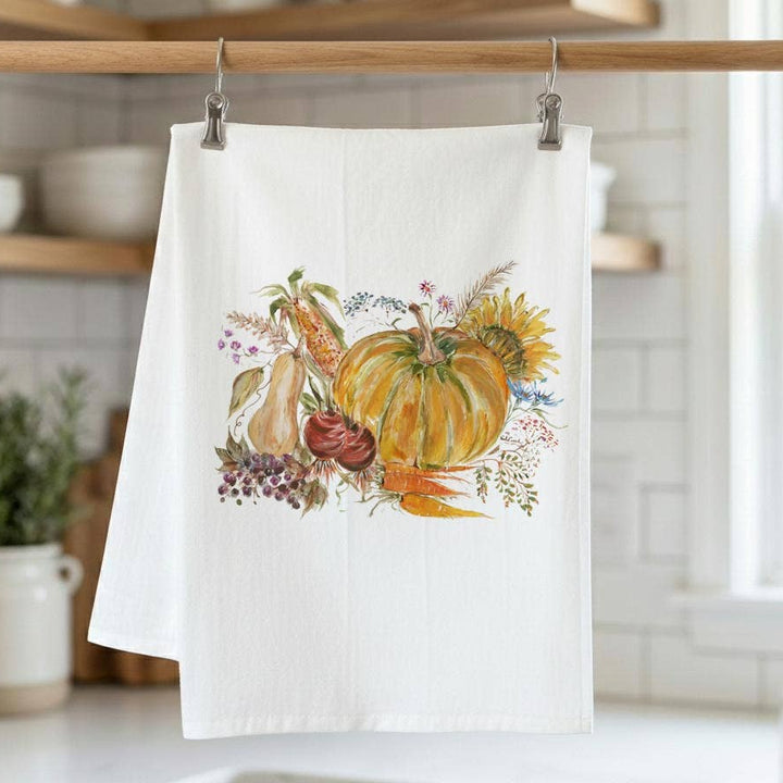 A white kitchen tea towel with a colorful autumn harvest illustration of pumpkins, gourds, flowers, and vegetables, hanging from a wooden rod in a bright kitchen.
